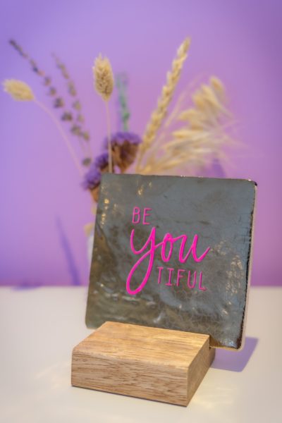 Decoratief bordje met de tekst “Be Youtiful” aan een paarse muur in de praktijkruimte van Zoëzo voor jou in Lutjebroek, met een bloemetje op de achtergrond.